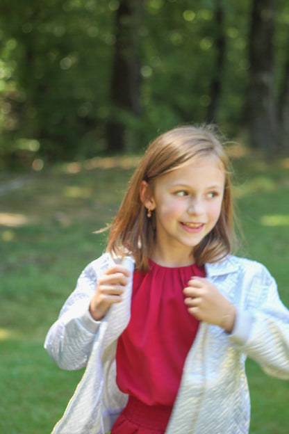 Delicate and dreamy, this silky dress is made just for girls who love a touch of elegance. It has soft pleats that gather gently at the neckline, creating a sweet, textured look. The bloused top flows beautifully over a fitted waist, introducing her to an appropriate and classic figure flattering look. Its comfy fabric makes it easy to move, twirl, and play—while still looking polished for special occasions or family celebrations.